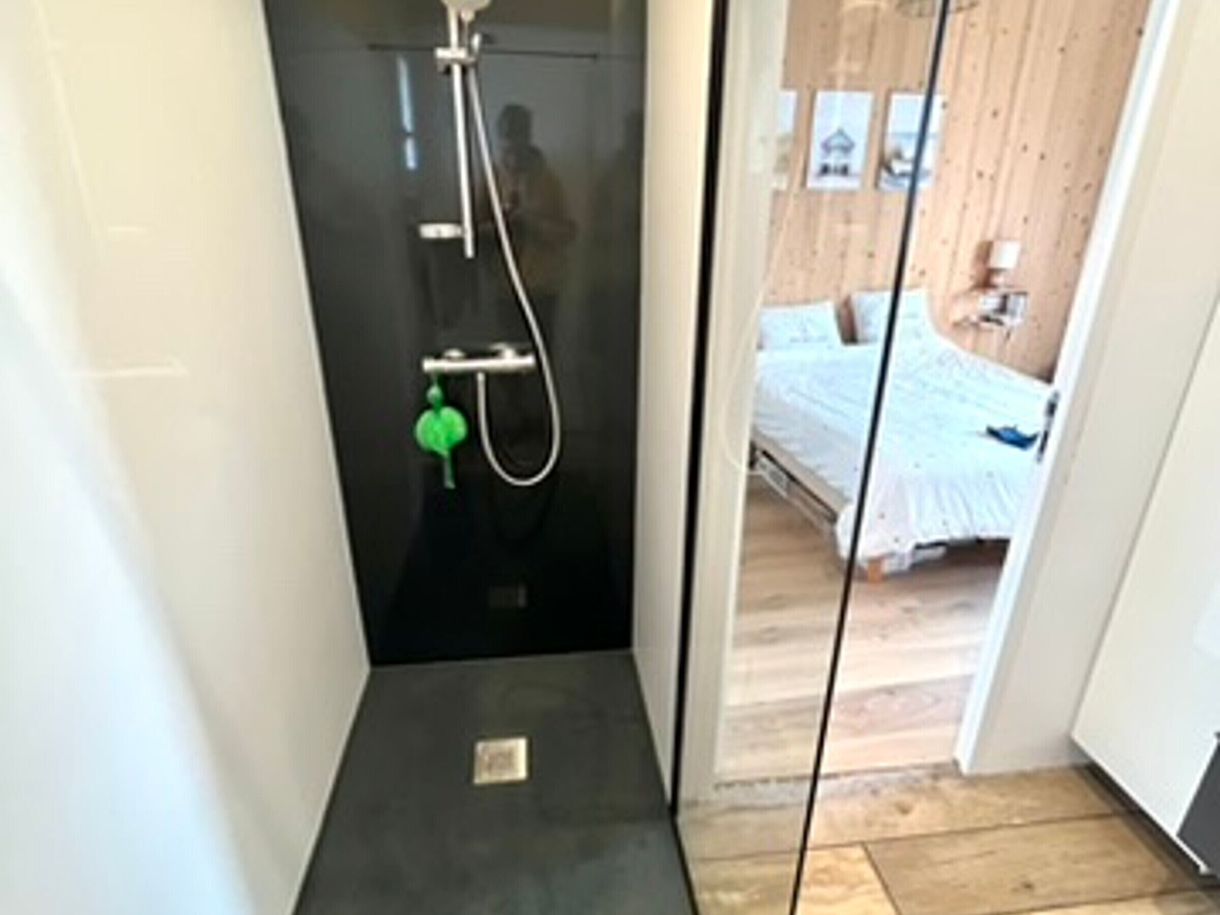 A narrow bathroom with a shower, a visible door leading to a bedroom with a bed and a nightstand.