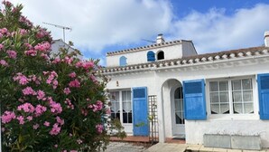 Un bâtiment blanc avec des volets bleus, un petit porche et un jardin où poussent des fleurs roses.
