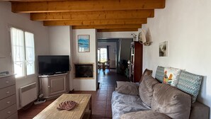Un salon avec un plafond en bois, une télévision, une cheminée, un canapé et, au fond, un coin repas.