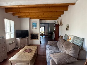 A living room with a wooden ceiling, a television, a fireplace, a sofa, and a dining area in the background. - House near sea and woods - 6 guests (La Guérinière)