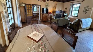 Een klassieke kamer met zichtbare houten balken, een open haard, een eethoek met tafel en stoelen, en een uitzicht op het landschap door een raam.