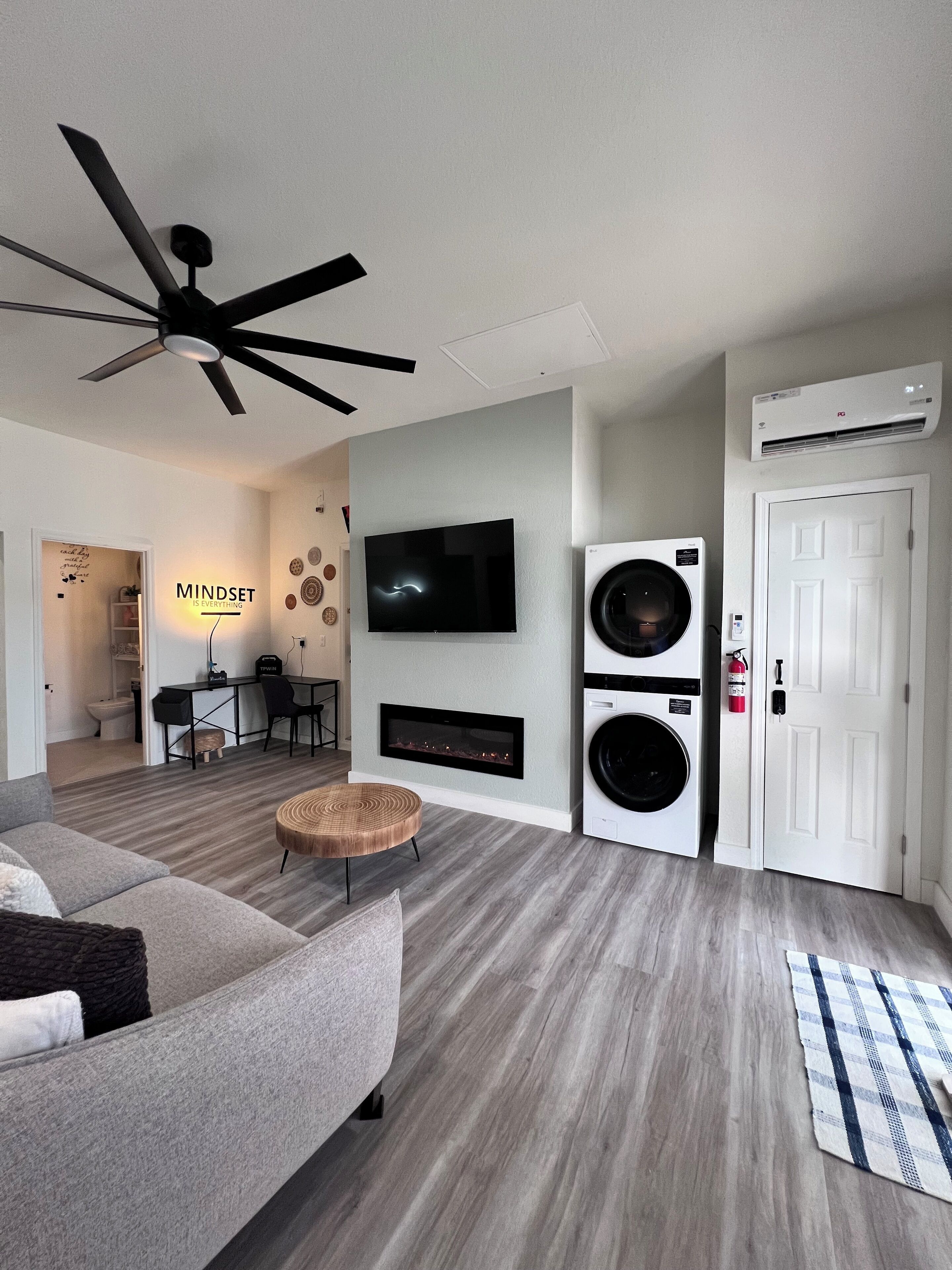 A modern living room with a ceiling fan, a flat-screen TV, a fireplace, a washing machine, and a door.