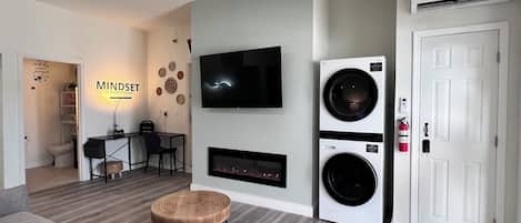A modern living room with a ceiling fan, a flat-screen TV, a fireplace, a washing machine, and a door.