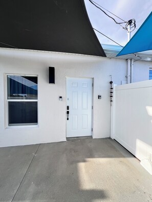 A white exterior wall with a white door, a window with a metal grill, and a blue awning. - Extended Stay Studio with Spa Rain Shower (Port St. Lucie)