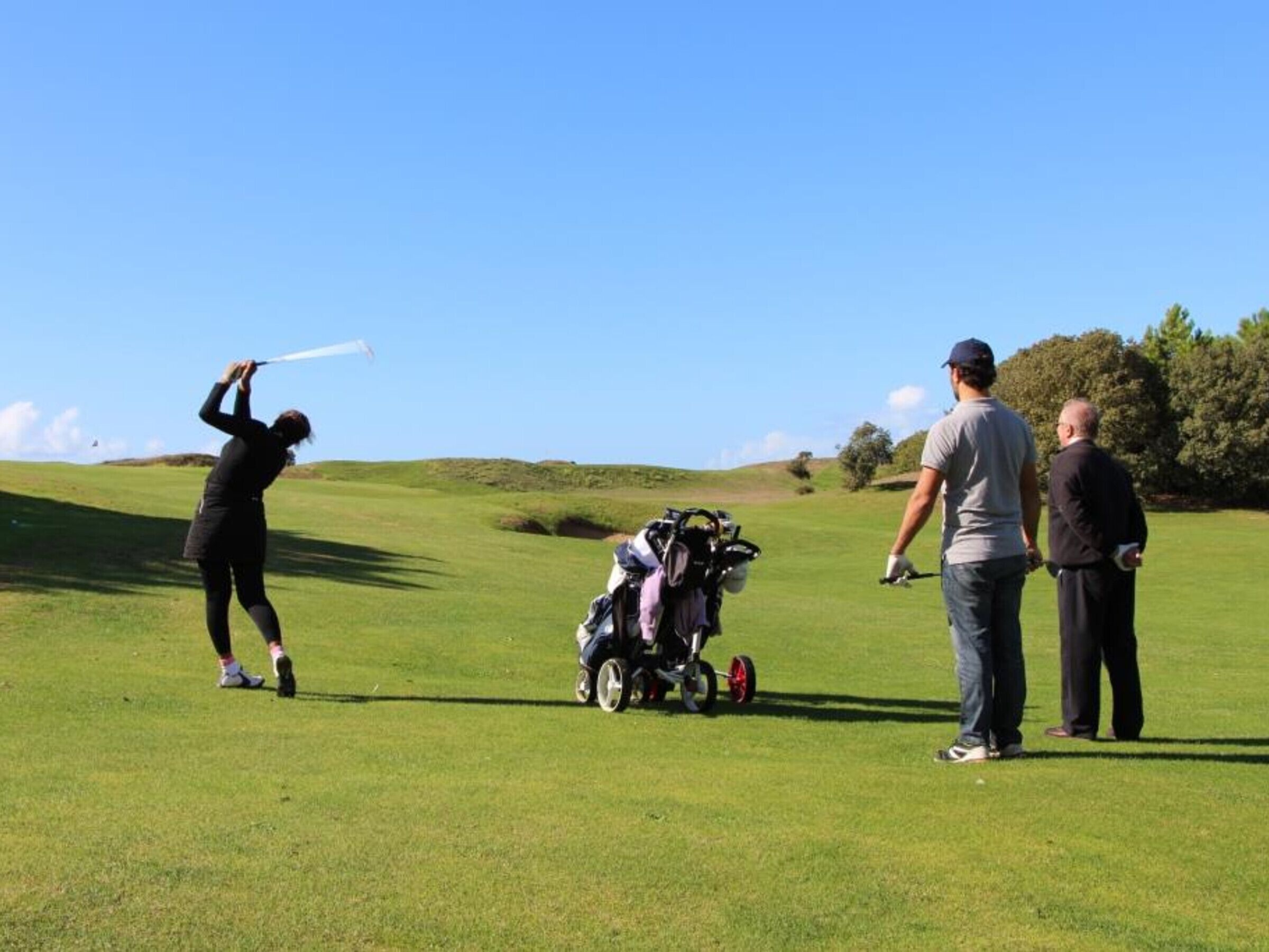 Ein Golfplatz mit einer Person, die bereit ist, einen Golfball zu schlagen, einer Golftasche und zwei weiteren Personen in der Nähe.