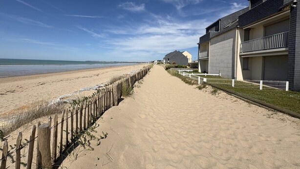 Un sentier de sable bordant la plage mène à une série de bâtiments modernes dotés de balcons, qui offrent une vue sur la mer.