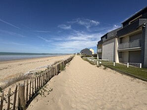Ein sandiger Küstenpfad führt zu einer Reihe moderner Gebäude mit Balkonen, mit Blick auf das Meer.