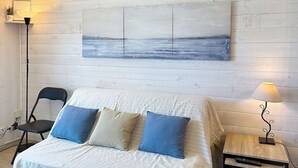 Un lit recouvert d’une couverture blanche, deux oreillers bleus, un tableau représentant un paysage marin, une chaise, une table de chevet avec une lampe et une télécommande.