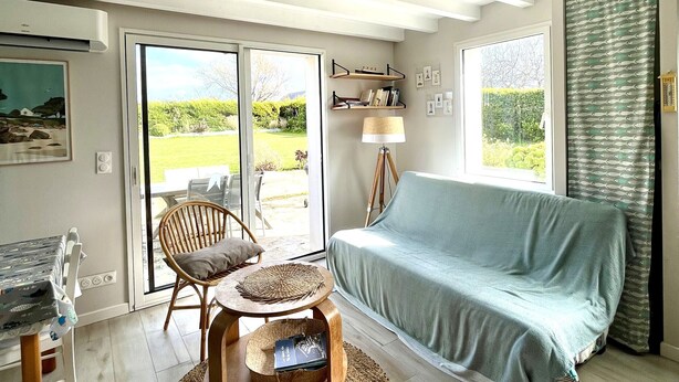 Ein Zimmer mit einer Couch, einem Sessel, einem Tisch, einer Lampe und Blick durch eine Glastür auf einen Garten.