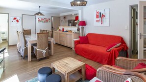 A modern living room with a red sofa, wooden dining table, wicker chairs, and a kitchen in the background. - Apartment near village - 10 guests (Barèges)