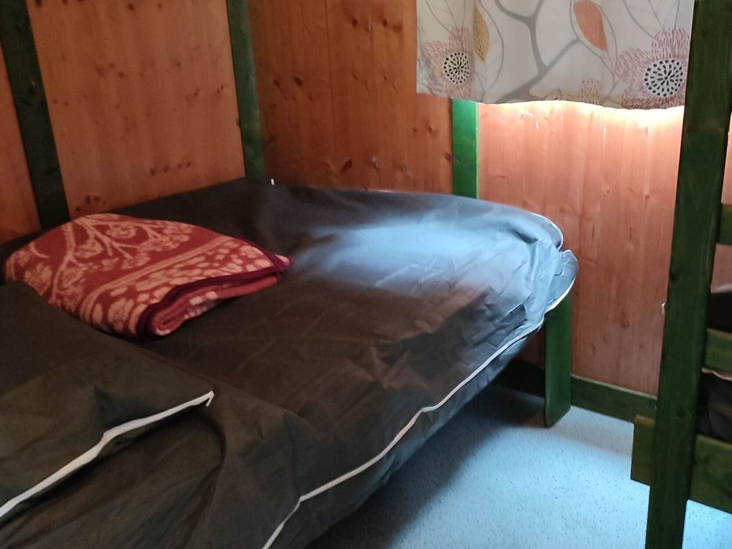 Un lit simple avec un matelas foncé et une couverture à motifs rouges, des lits superposés en bois avec des touches de vert, et une décoration murale florale.