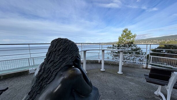 En skulptur som föreställer en kvinna som sitter på en bänk och blickar ut över ett havsnära landskap.