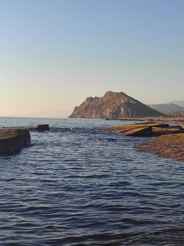 Un paysage côtier caractérisé par une colline rocheuse imposante, une mer calme et une plage de galets.