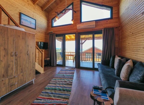 A wooden cabin interior with a sofa, TV, and a staircase leading to a balcony. - Koru Wooden House (Gazipasa)