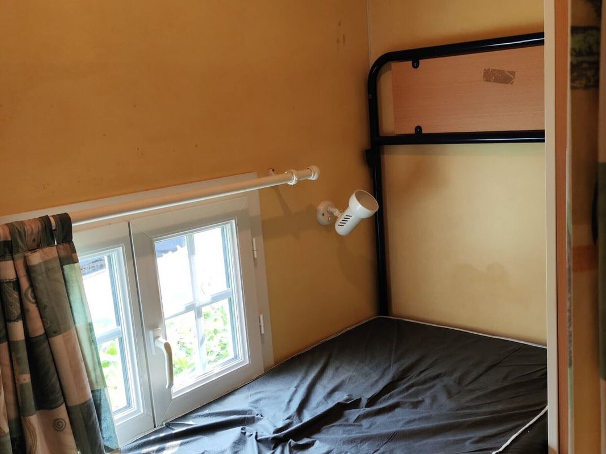 Een eenpersoonsbed met hoofdeinde, een raam met gordijnen en een witte beker aan de muur.