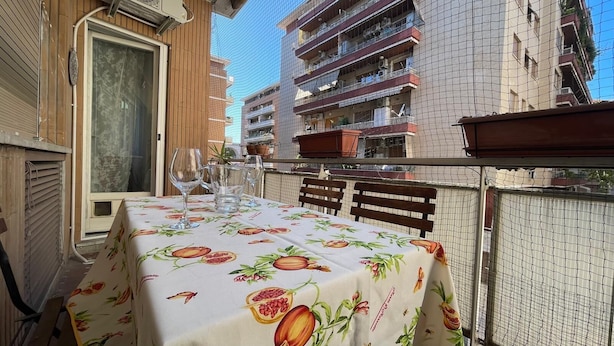 A table with a fruit pattern tablecloth, a window with blinds, and a view of a multi-story building. - Livia's Vatican House - Metro Aurelia (Rome)