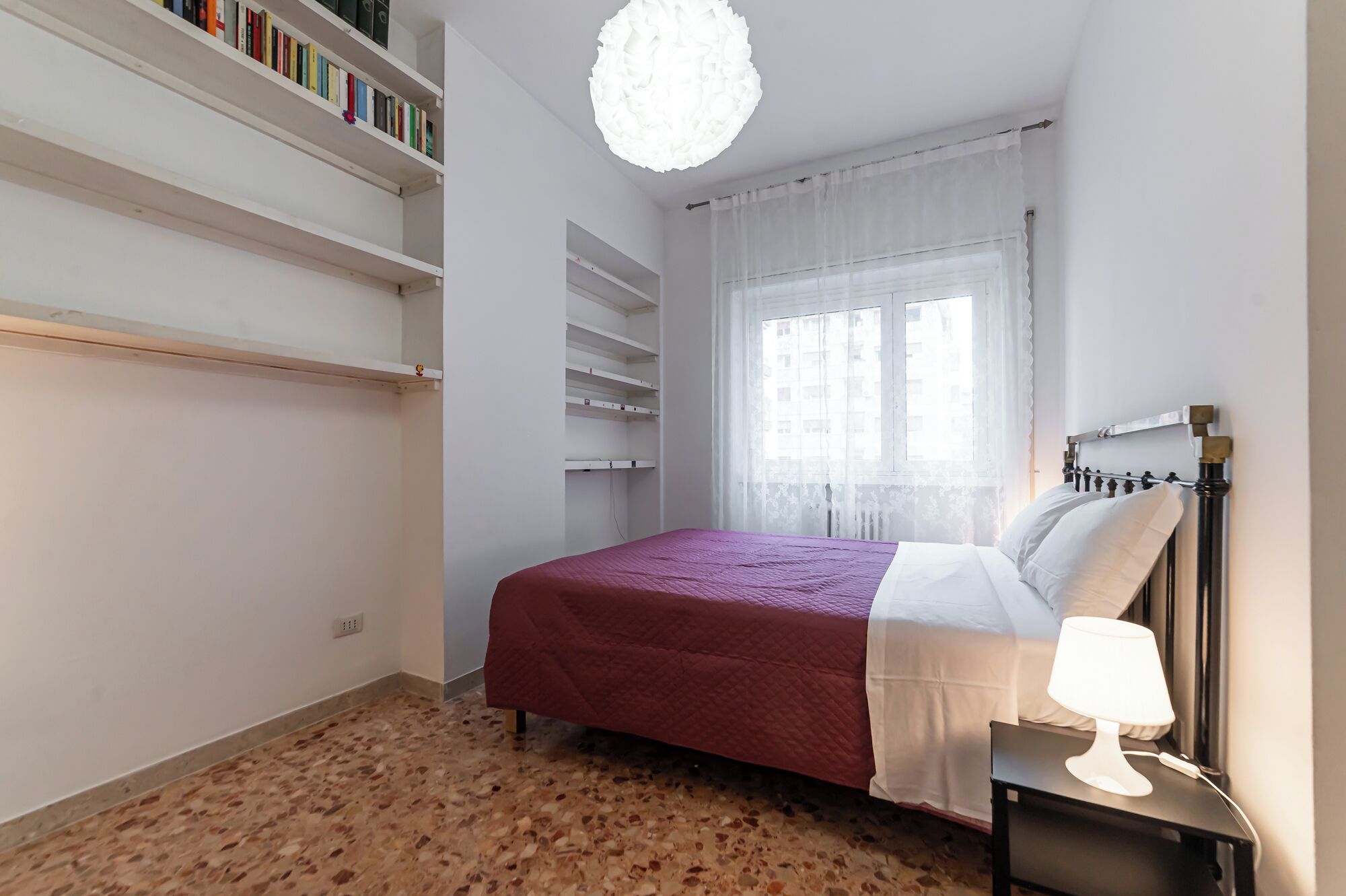 A bedroom with a bed, a nightstand, a lamp, a window with sheer curtains, and a bookshelf.