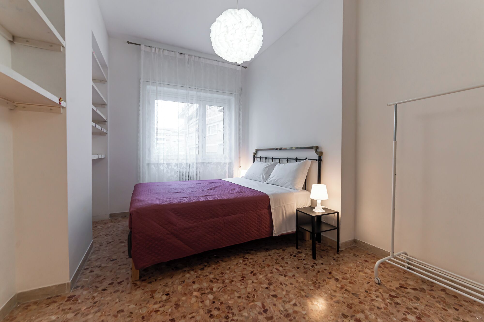 A bedroom with a bed, a nightstand, a lamp, a window with sheer curtains, a ceiling light, and a radiator.