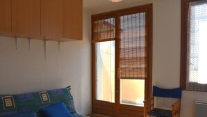 A room with a sofa, a small table, a chair, a sliding door with a view, and a window with a curtain.