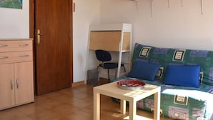 A room with a wooden door, a desk with a chair, a bed with patterned bedding, and a small table with a tray.