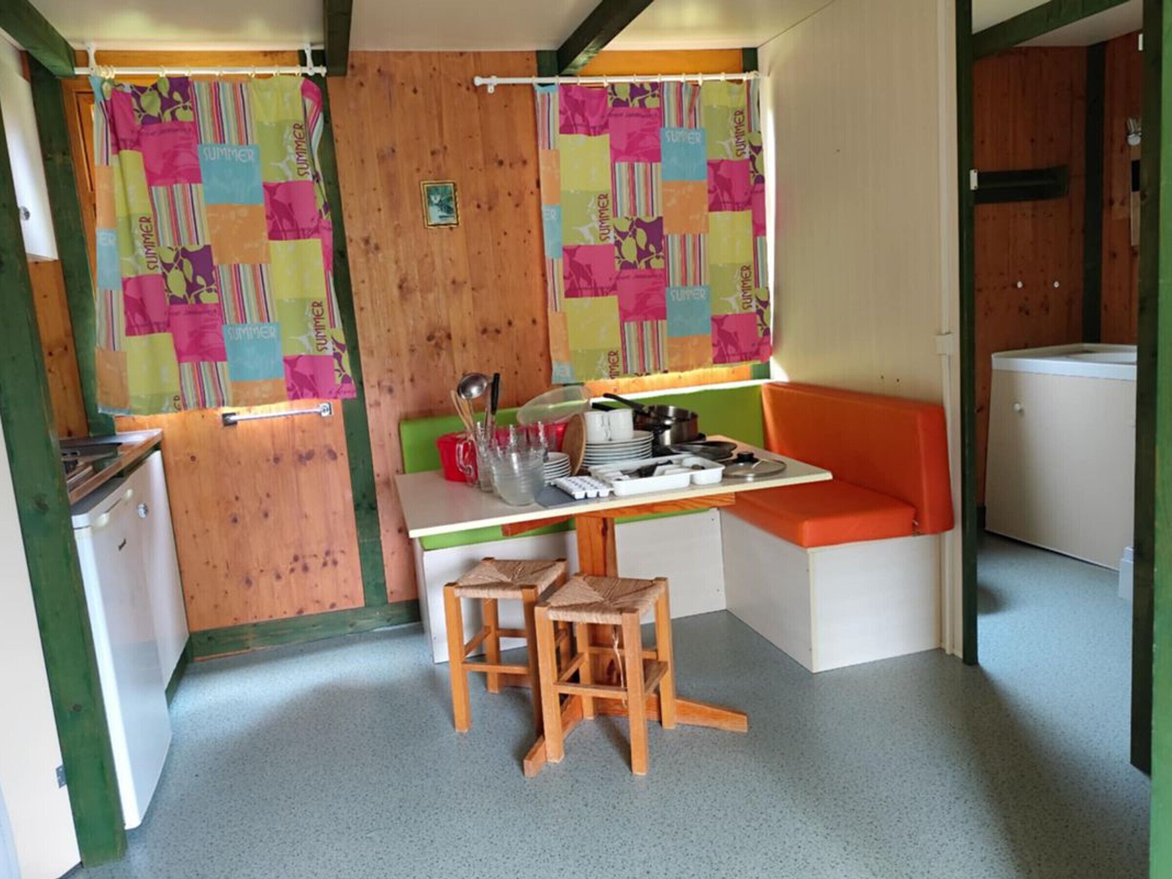 A compact kitchen with a wooden table, two stools, a bench, and a refrigerator.