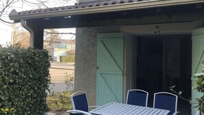 Ein überdachter Außenbereich mit Tisch und Stühlen, einer grünen Tür und einer Hecke.