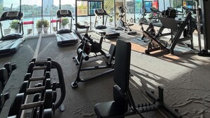 Une salle de sport équipée de tapis de course, de vélos elliptiques et d’appareils de musculation, avec de grandes fenêtres et une vue sur les bâtiments.