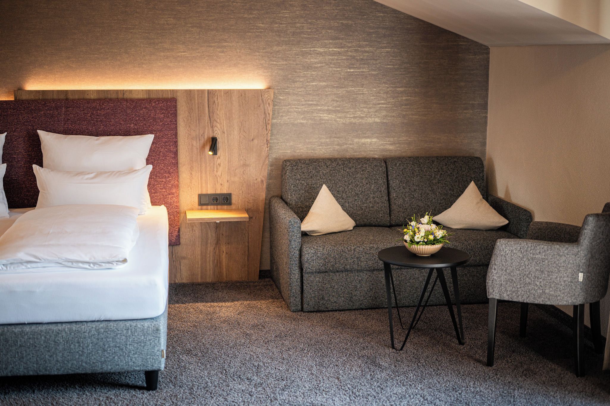 Ein Hotelzimmer mit einem Bett, einer Couch, einem Sessel, einem kleinen Tisch mit Blumen und einer Wandleuchte.