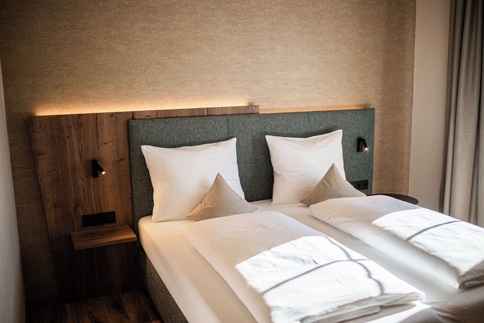 A double bed with white linens, two pillows, a headboard with a textured fabric, and a bedside table with a lamp.