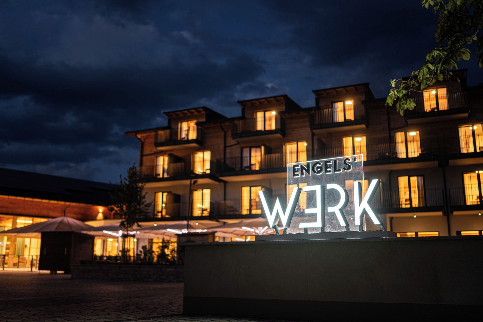 Entrada de un hotel con un cartel que dice 'ENGELS' WERK' y un edificio con múltiples ventanas iluminadas en la noche.