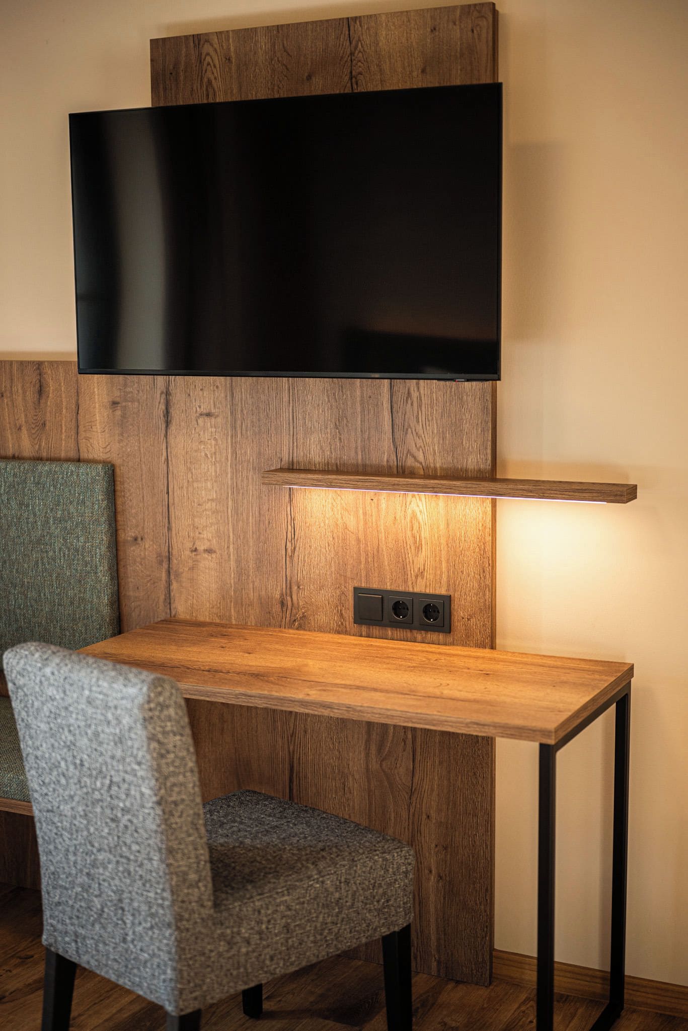 A modern room with a wooden desk, a chair, a flat-screen TV mounted on the wall, and a built-in shelf with a light underneath.