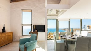 A room with a large window, a television, a wooden cabinet, a blue armchair, and a dining area with a table set for two. - Villas Maya 3-bedroom villa in fabulous Chorafakia with private pool (Chorafakia)