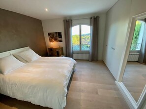 Ein Schlafzimmer mit einem großen Bett, einem Nachttisch, einem Fenster mit Vorhängen und Blick ins Grüne.