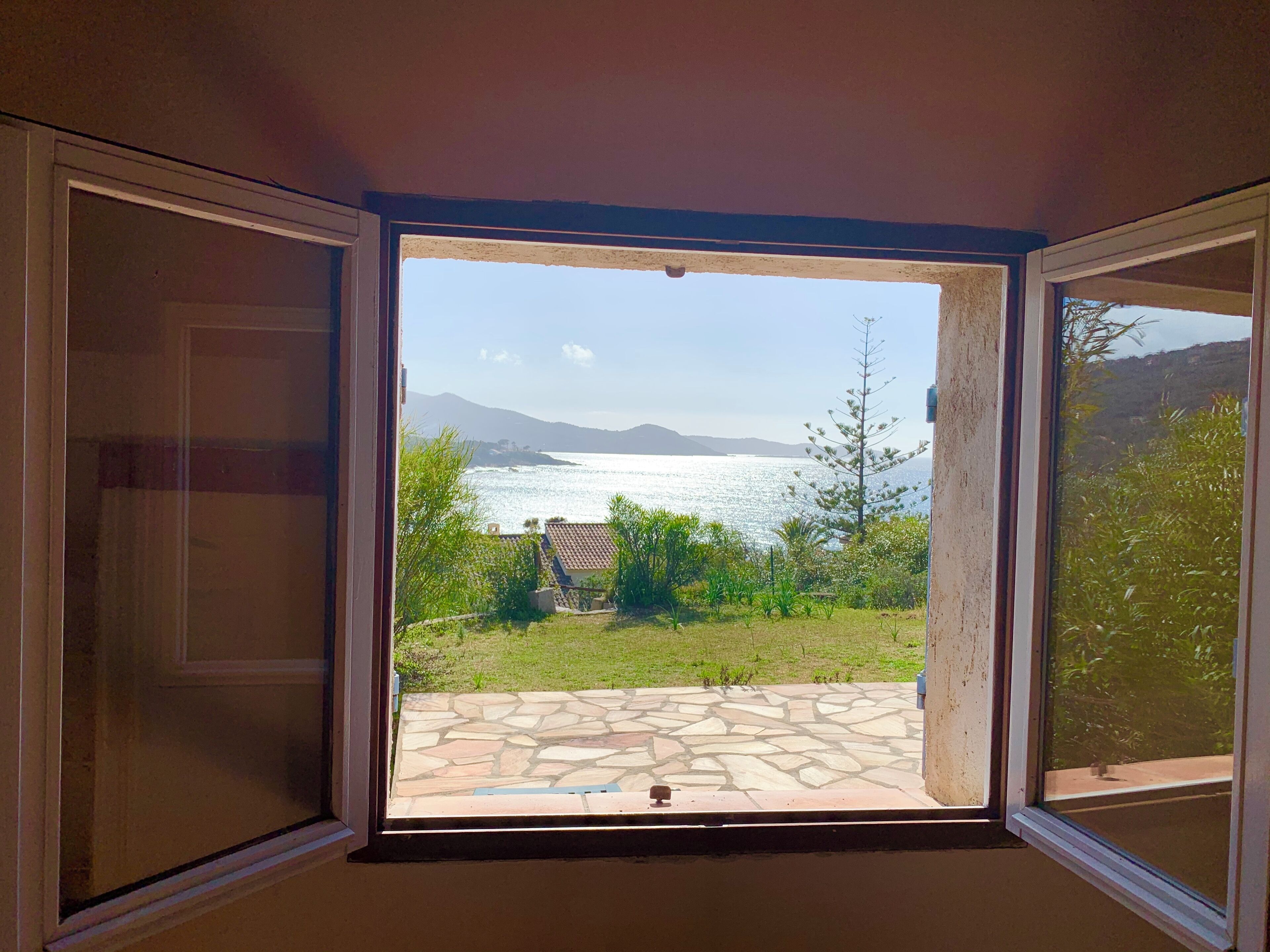 An open window with a view of a stone patio, a grassy area, and a body of water with mountains in the distance.