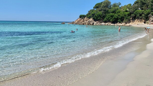 A beach with clear water, pebbly shore, and a rocky cliff with trees.