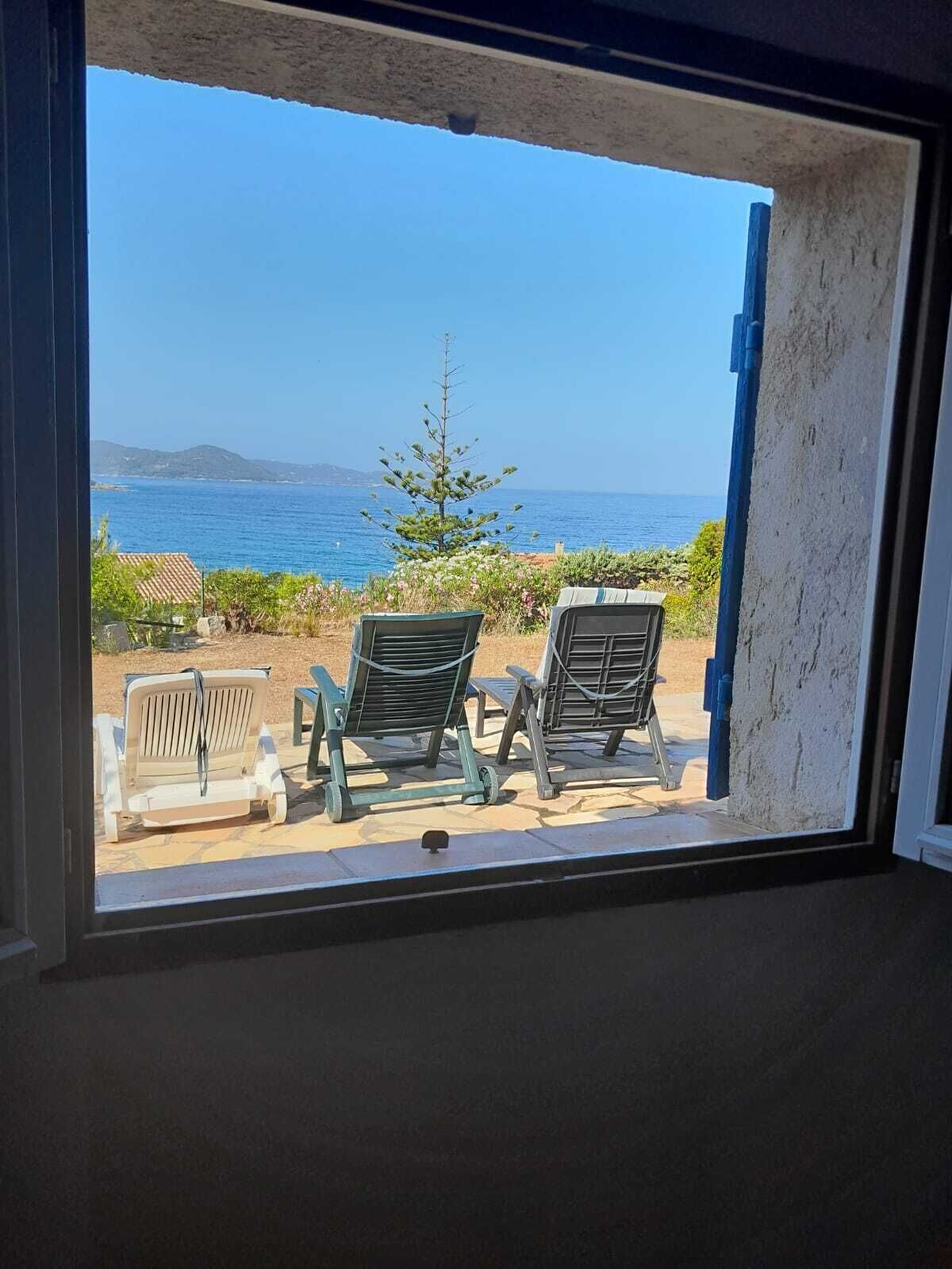 Une vue depuis une fenêtre sur une zone côtière, avec deux chaises longues et un petit arbre, donnant sur la mer et les collines lointaines.