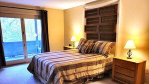 Quarto com cama, mesa de cabeceira, luminária, janela com cortinas e vista para o exterior.