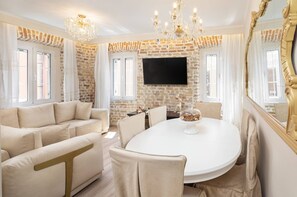 A dining area with a brick wall, a flat-screen TV, a chandelier, and a round table with chairs. - Borgo Veneziano by Corfuescapes (Corfu)