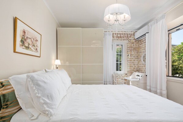 A bedroom with a large bed, a chandelier, a window with curtains, a radiator, and a painting on the wall. - Borgo Veneziano by Corfuescapes (Corfu)