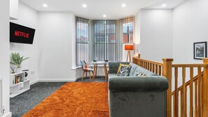 Een moderne woonkamer met een grijze bank, oranje vloerkleed, een aan de muur bevestigde televisie en een trap.