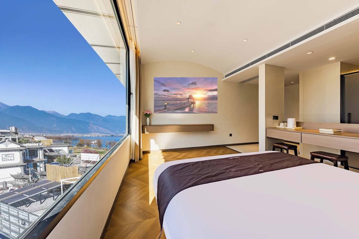 Une chambre d’hôtel avec un grand lit, un bureau, une chaise, une fenêtre donnant sur la ville et un tableau représentant un coucher de soleil accroché au mur.