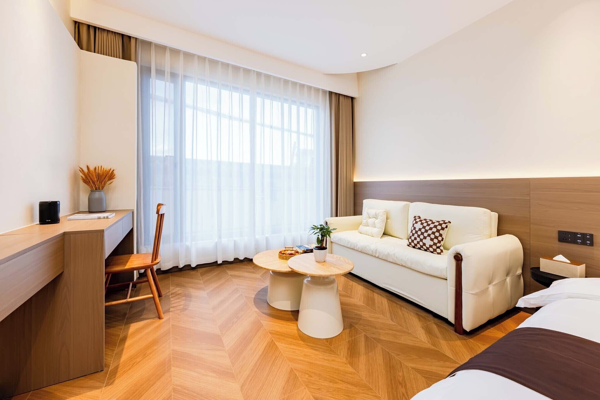 A modern hotel room with a wooden herringbone floor, a white sofa with a patterned pillow, a small round table, a desk with a chair, and a large window with sheer curtains.