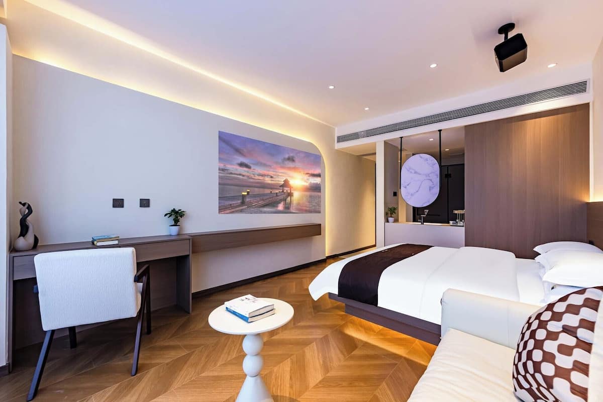 Une chambre d’hôtel moderne avec un parquet en bois disposé en motif chevrons, un lit avec une literie blanche, un bureau avec une chaise, une petite table ronde avec un livre, et un tableau représentant un coucher de soleil accroché au mur.