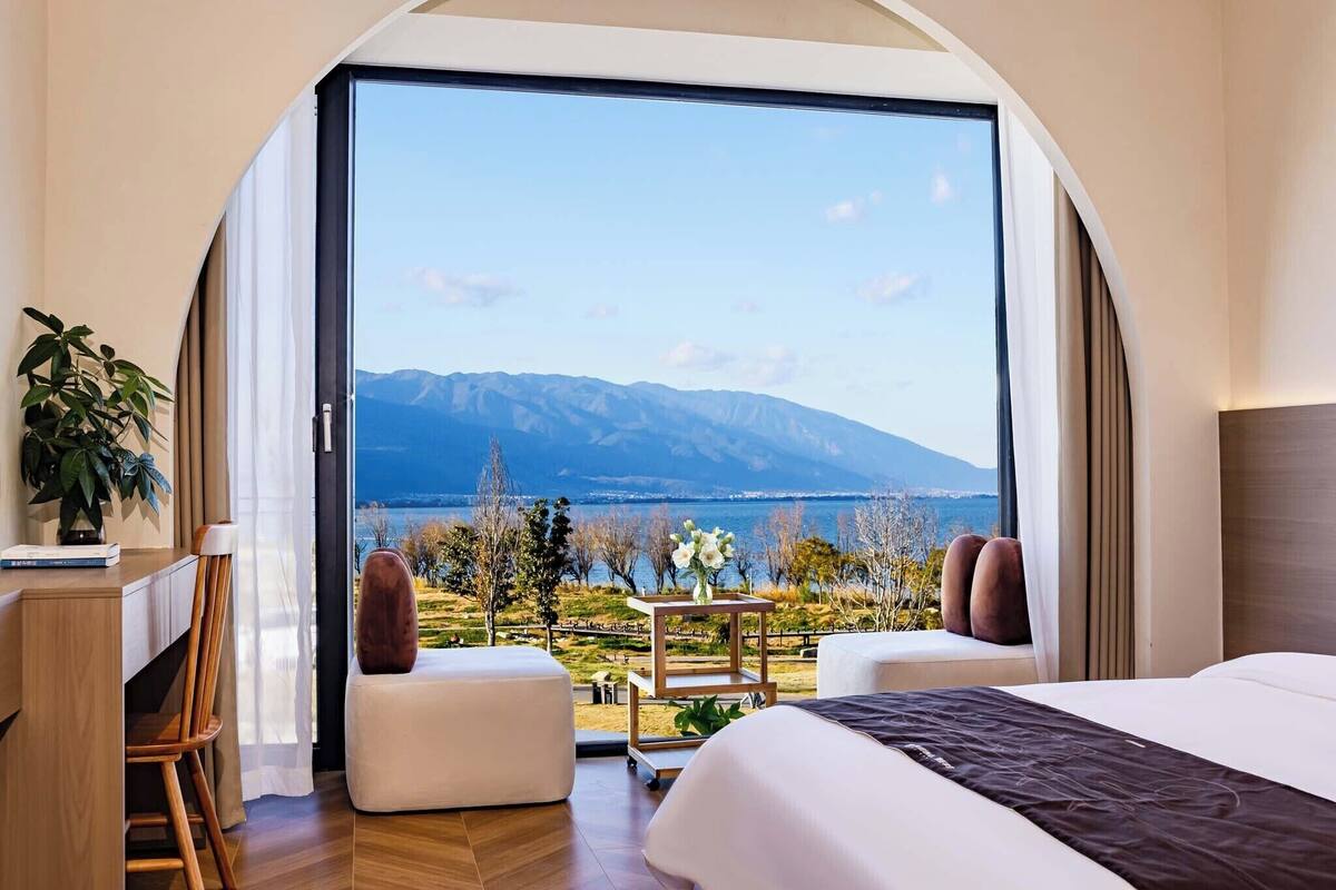 Une chambre avec une grande fenêtre, un bureau, deux poufs et un lit.