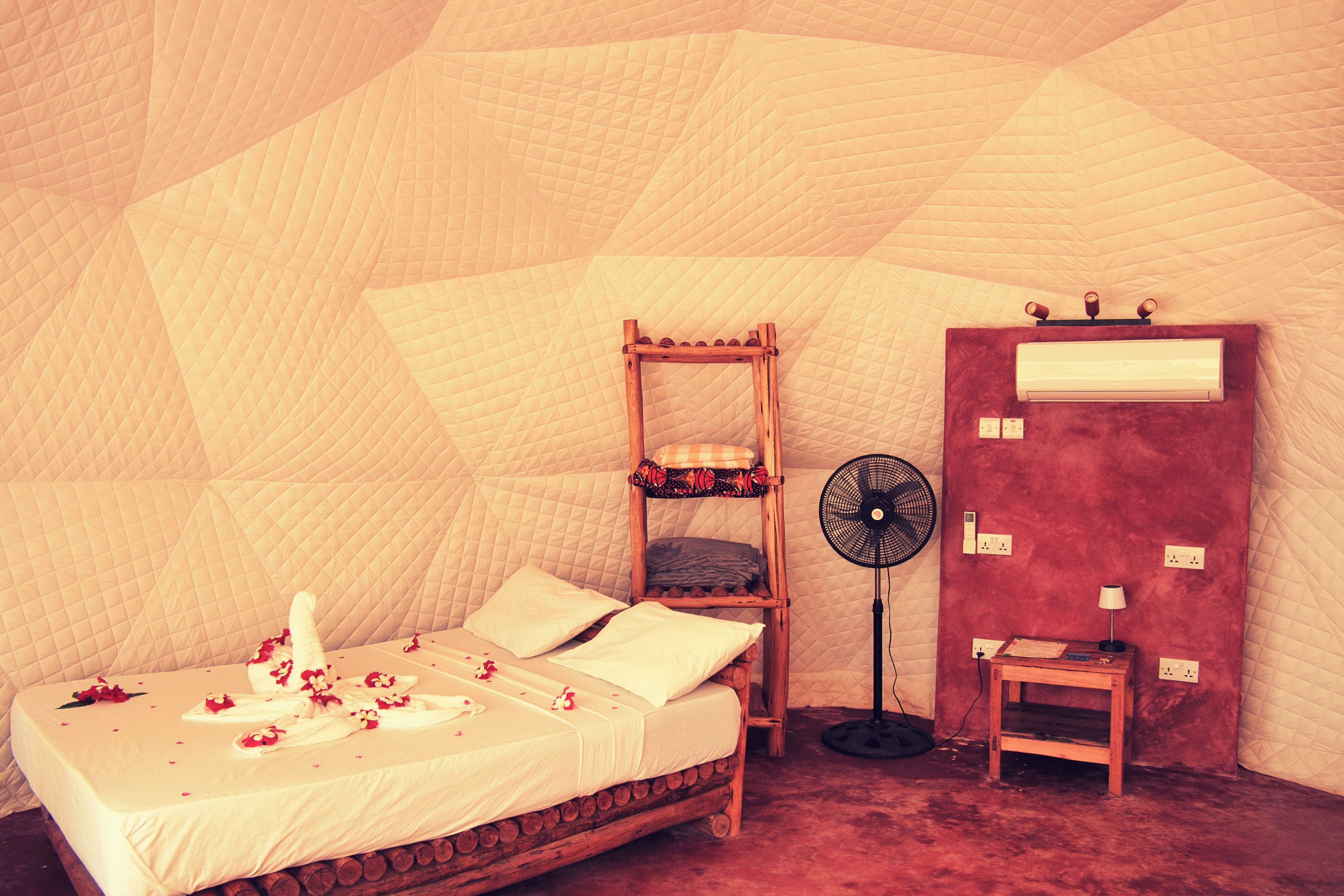 Een kamer met een bed, een houten nachtkastje, een ventilator en een rode muur met stopcontacten.