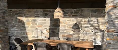 Un espace extérieur aménagé pour les repas, avec des murs en pierre, une table et des chaises en bois, un luminaire suspendu et un banc en pierre.