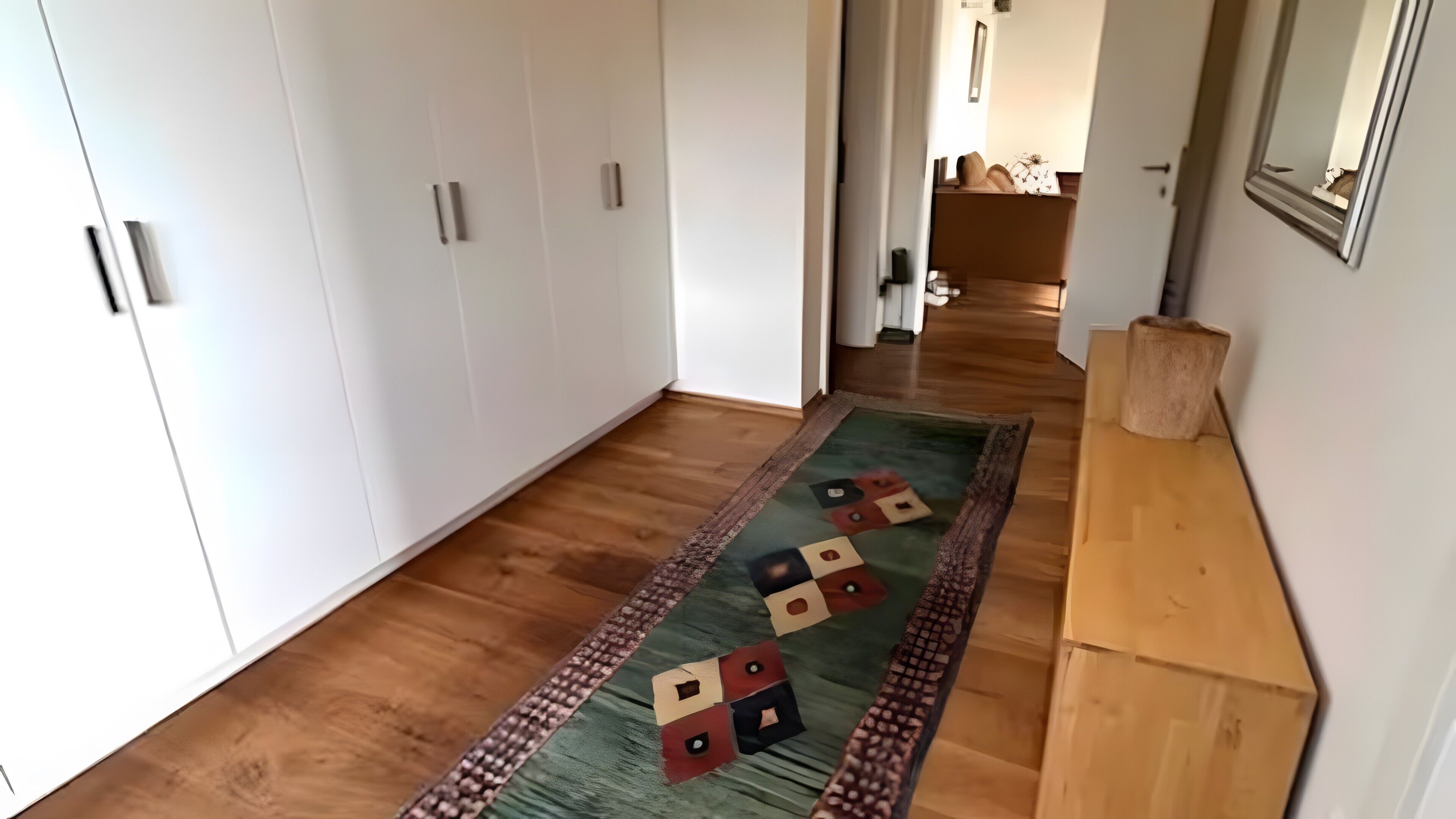 Käytävän lattia on parkettia, siellä on vihreä-punainen kuvioitu matto ja puinen konsolipöytä.