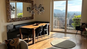A modern living room with a wooden dining table, black chairs, a black sofa, a large mirror, and a view of a cityscape through a sliding glass door.