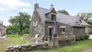 A stone house with a sloped roof, a wooden fence, and a garden area with a chair and a table. - Gîte La P'tite Longère – Independent Cottage in a Peaceful Countryside Setting (Saint-Jacut-les-Pins)