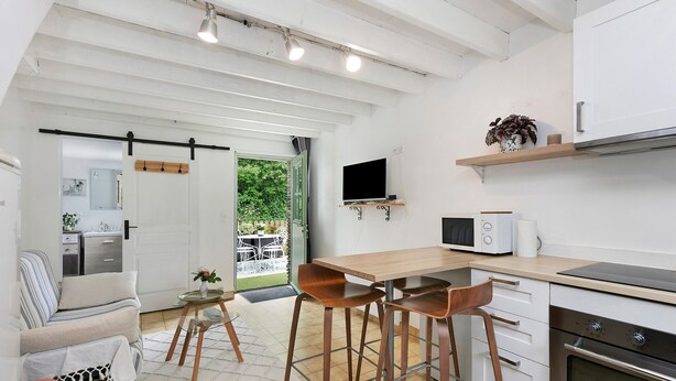 A modern kitchen with a dining area, a TV mounted above the counter, and a sliding door leading to an outdoor patio. - Gîte La P'tite Longère – Independent Cottage in a Peaceful Countryside Setting (Saint-Jacut-les-Pins)
